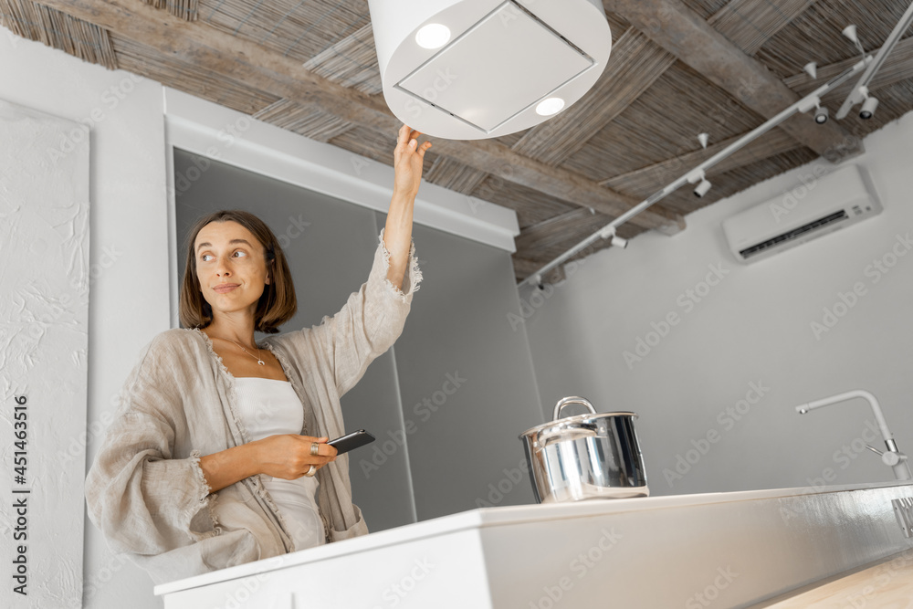 Wall mural Young woman controls hood, while standing with cell phone and cooking at modern kitchen. Concept of smart technologies of kitchen appliances