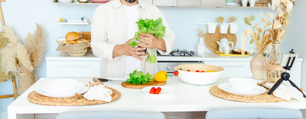 happy young, handsome, bearded man is standing in the modern kitchen prepares a salad of fresh vegetables, cuts vegetables with a knife on a cutting board, cooking as concept of a man's hobby