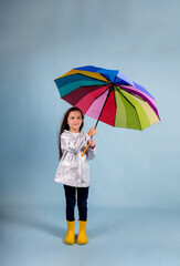 a little girl in a raincoat and yellow rubber boots stands and holds a multi-colored umbrella on a blue background with a place for text