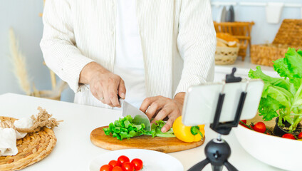 Obraz na płótnie Canvas happy young, handsome, bearded man is standing in the modern kitchen prepares a salad of fresh vegetables lettuce, tomatoes, sweet pepper with a knife on a cutting board, records video for food blog