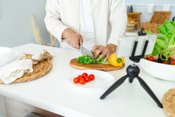 happy young, handsome, bearded man is standing in the modern kitchen prepares a salad of fresh vegetables lettuce, tomatoes, sweet pepper with a knife on a cutting board, records video for food blog