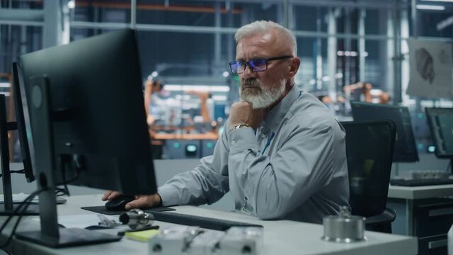 Car Factory Office: Portrait of Senior Male Chief Engineer Working on Desktop Computer in Automated Robot Arm Assembly Line Manufacturing High-Tech Electric Vehicles. Electronics Production Conveyor