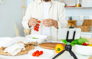 happy young, handsome, bearded man is standing in the modern kitchen prepares a salad of fresh vegetables lettuce, tomatoes, sweet pepper with a knife on a cutting board, records video for food blog