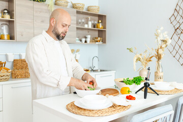 happy young, handsome, bearded man is standing in the modern kitchen prepares a salad of fresh vegetables lettuce, tomatoes, sweet pepper with a knife on a cutting board, records video for food blog