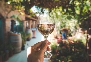 Fototapeta premium Glass of wine in hand. A glass of young fresh rose wine against the backdrop of a summer cafe in a Mediterranean seaside tourist town in the summer under sunlight. Summer, travel, lifestyle