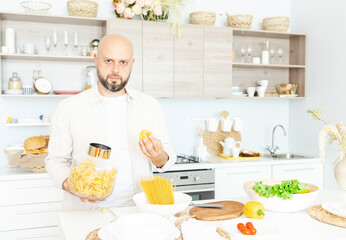 happy young, handsome, bearded man is standing in the modern kitchen with pasta in his hands and vegetables on table, cooking as concept of a man's hobby