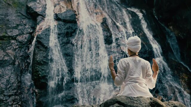 Young woman in white clothes doing yoga on the rocks against the background of a waterfall
