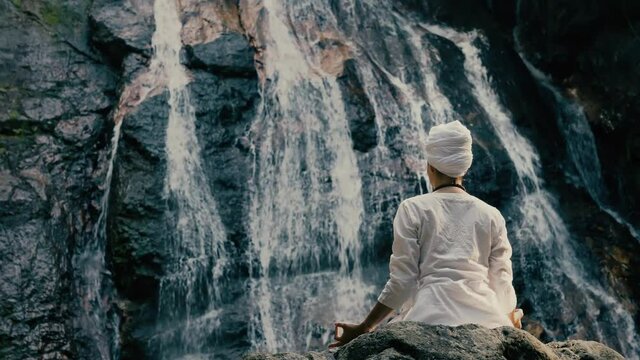 Young woman in white clothes doing yoga on the rocks against the background of a waterfall