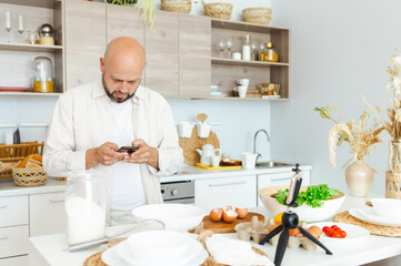 happy young, handsome, bearded man is standing in modern kitchen and talking on phone before prepares a salad of fresh vegetables lettuce, tomatoes, sweet pepper and eggs, concept of culinary blog