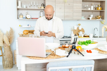 happy young, handsome, bearded man is standing in modern kitchen prepares salad fresh vegetables lettuce, tomatoes, sweet pepper with knife on a cutting board, records video for food blog with laptop