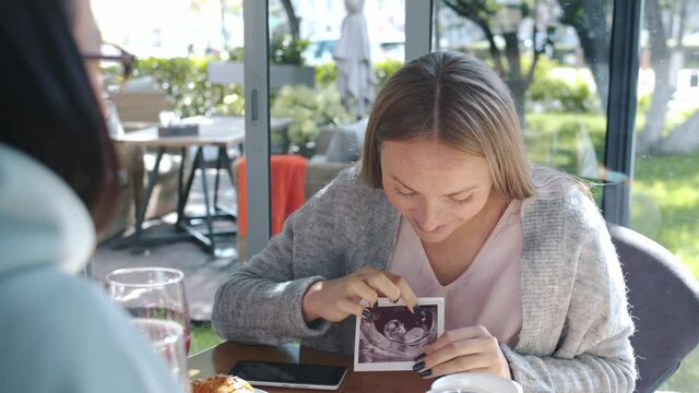 Future mother cheerful young lady is showing ultrasound image of baby to friends in cafe sharing emotions. Pregnancy and leisure time concept.