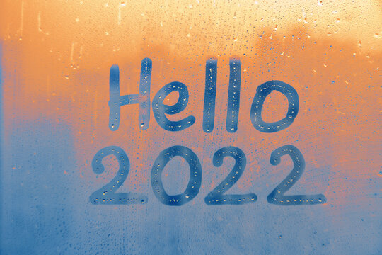 lttering Hello and numbers 2022 of new year paint with finger on splashed by rain foggy glass on blue and sunset window