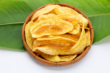 Banana slice chips in bamboo basket on green leaf and white background.