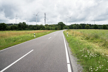 Deserted country road through a meadow with wind turbines in background on a cloudy summer day