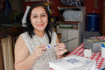 Happy seniors hispanic woman doing a craft at home-woman smiling at camera as she paints with...