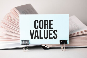 A white card with the text core values stands on a clip for papers on the table against the background of books. Defocus