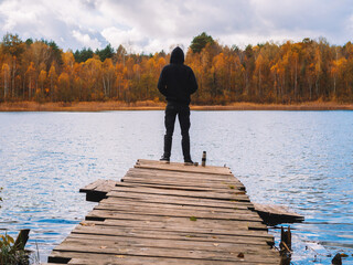 Rear view, a man on a wooden bridge by the lake against the background of an autumn forest.