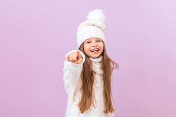 A child in winter clothes points to the space in front of him and smiles against an isolated pink background.