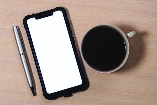 Mockup image of a smartphone with the blank white screen, pen and a cup of coffee on a wooden table.