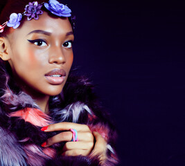 young pretty african american woman in spotted fur coat and flowers on head smiling on black background, fashion people concept