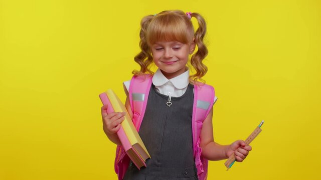 Cheerful funny teenage schoolgirl kid with books dressed in uniform wears pink backpack smiling, looking at camera isolated on yellow studio background. Teenage child education, back to school, study
