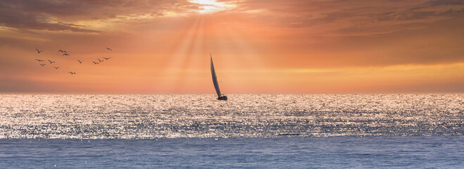 Sailboat sailing towards the horizon at a warm orange cloudy sunset