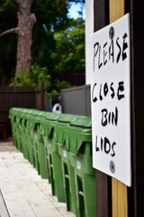Row of green trash bins in the community with a sign that instructs Please Close Bin Lids