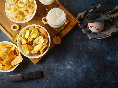 Beer in glasses, chips, snacks, onion rings, baseball glove and ball, remote control. High angle view. Rest with friends. Watching a baseball game on TV. There are no people in the photo.
