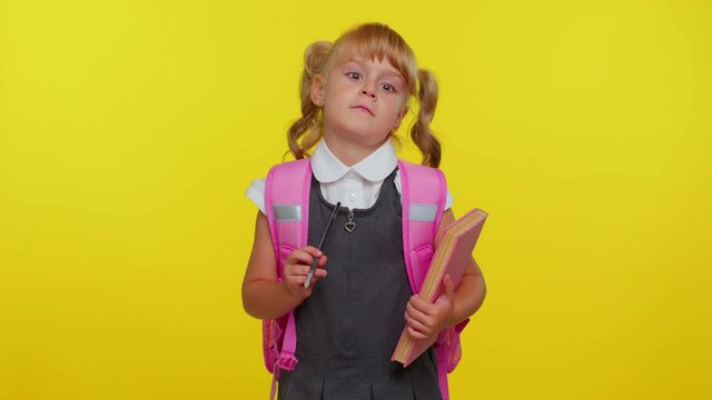 Smiling funny schoolgirl wears backpack, making humorous face with eyes crossed, playing fool, having fun with stupid brainless dumb expression over yellow studio background. Back to school concept