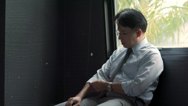 Stressed Asian businessman sitting by the window holding financial bill thinking of debt and business problem. Unemployed man worry about money management. Mental health and economic problem concept