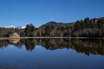 Obraz premium The lake house in the forest. Beautiful view of the summer cabin, calm lake, mountains and forest under a blue sky. The perfect reflection in the water surface. 