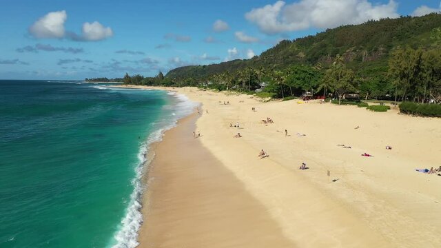 Aerial drone footage above a beach on Oahu, Hawaii, USA. Broad, sandy beach with turquoise water and white waves, and sunbathers visible on the sand. Green forest to the right of the beach.