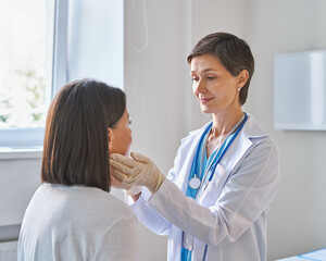 Friendly middle-aged woman doctor wearing gloves checking sore throat or thyroid glands, touching...
