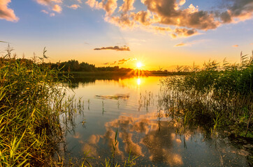 Fototapeta na wymiar Scenic view at beautiful summer sunset on lake with reflection on water with reeds, grass, golden sun rays, calm water ,deep blue cloudy sky and glow on a background, spring evening landscape