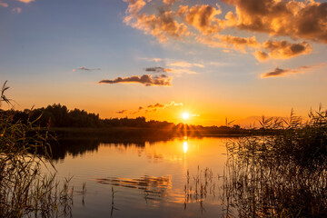 Scenic view at beautiful summer sunset on lake with reflection on water with reeds, grass, golden sun rays, calm water ,deep blue cloudy sky and glow on a background, spring evening landscape