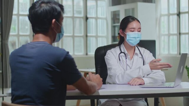Asian Woman Doctor Is Talking With Young Male Patient During Consultation In A Health Clinic. Both Wear Face Masks. Physician In Lab Coat Sitting Behind A Computer Desk In Hospital Office
