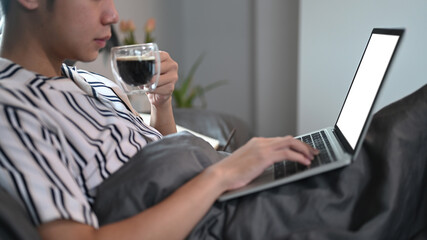 Young man drinking coffee and using computer laptop on a bed.