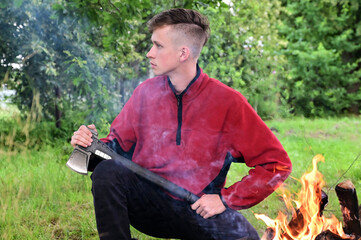 young serious man with a mohawk hairstyle is sitting with an axe by a fire in the forest. Take a break. Path to nature. Concept of adventure