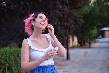 Young caucasian woman with pink hair putting wireless earbuds in a park.