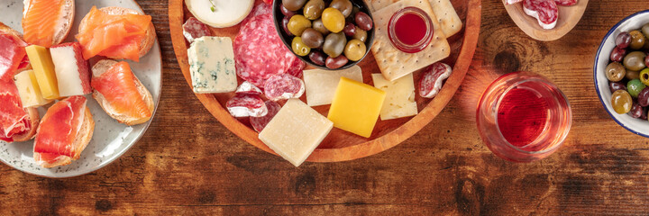 Charcuterie and cheese board panorama with rose wine, olives, and a place for text, shot from above on a rustic background. Spanish tapas or Italian antipasti. Salmon sandwiches, blue cheese etc