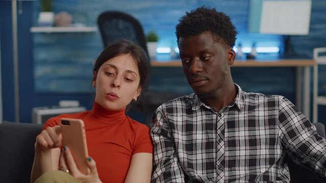 Modern interracial couple using smartphone in living room. Young multi ethnic people looking at digital device and relaxing on couch at home. Cheerful husband and wife sitting together