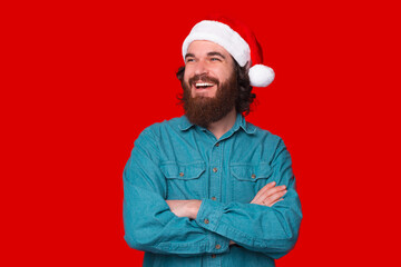 Photo of smiling bearded guy wearing santa claus hat, with folded hands and looking away over red background