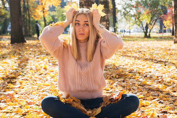 Young pretty woman is sitting on tground in autumn park with leaves in her hands