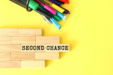 Wooden blocks stacked next to pens and pencils on a yellow background. SECOND CHANCE text on a wooden block.