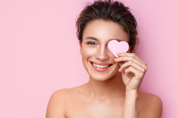 Obraz na płótnie Canvas Smiling woman with cosmetic sponge covering one eye. Photo of woman with perfect makeup on pink background. Beauty concept