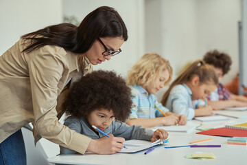 Attentive young female teacher helping little schoolboy. Kids sitting at the table, studying in elementary school classroom