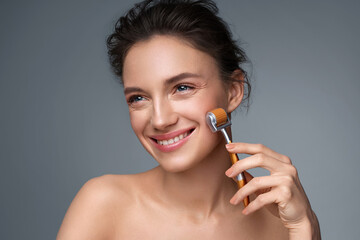 Beautiful woman using derma roller for her facial skin. Photo of woman on gray background. Beauty and skin care concept - obrazy, fototapety, plakaty