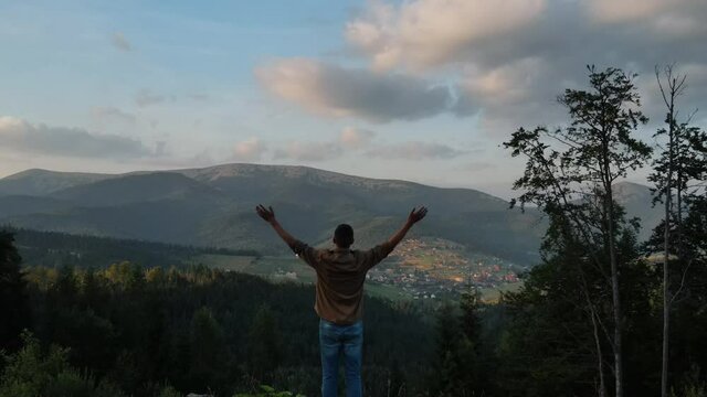 Aerial drone follows of young man running on green field on background mountains summit during sunset or sunrise, raises arms into air, happy and drunk on life, youth and happiness. Travel concept