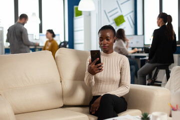 African manager woman discussing with remote colleagues on video call holding smartphone using headphones sitting on couch in business modern office. Diverse coworkers planning new financial project