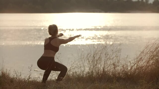 Fit Sporty Woman Squats Exercising At Sunset. Stretching Fitness Workout Activity. Squats Workout Practicing. Sport Recreation Healthy Lifestyle. Warming Up Aerobic Exercises. Cardio Squat Training.